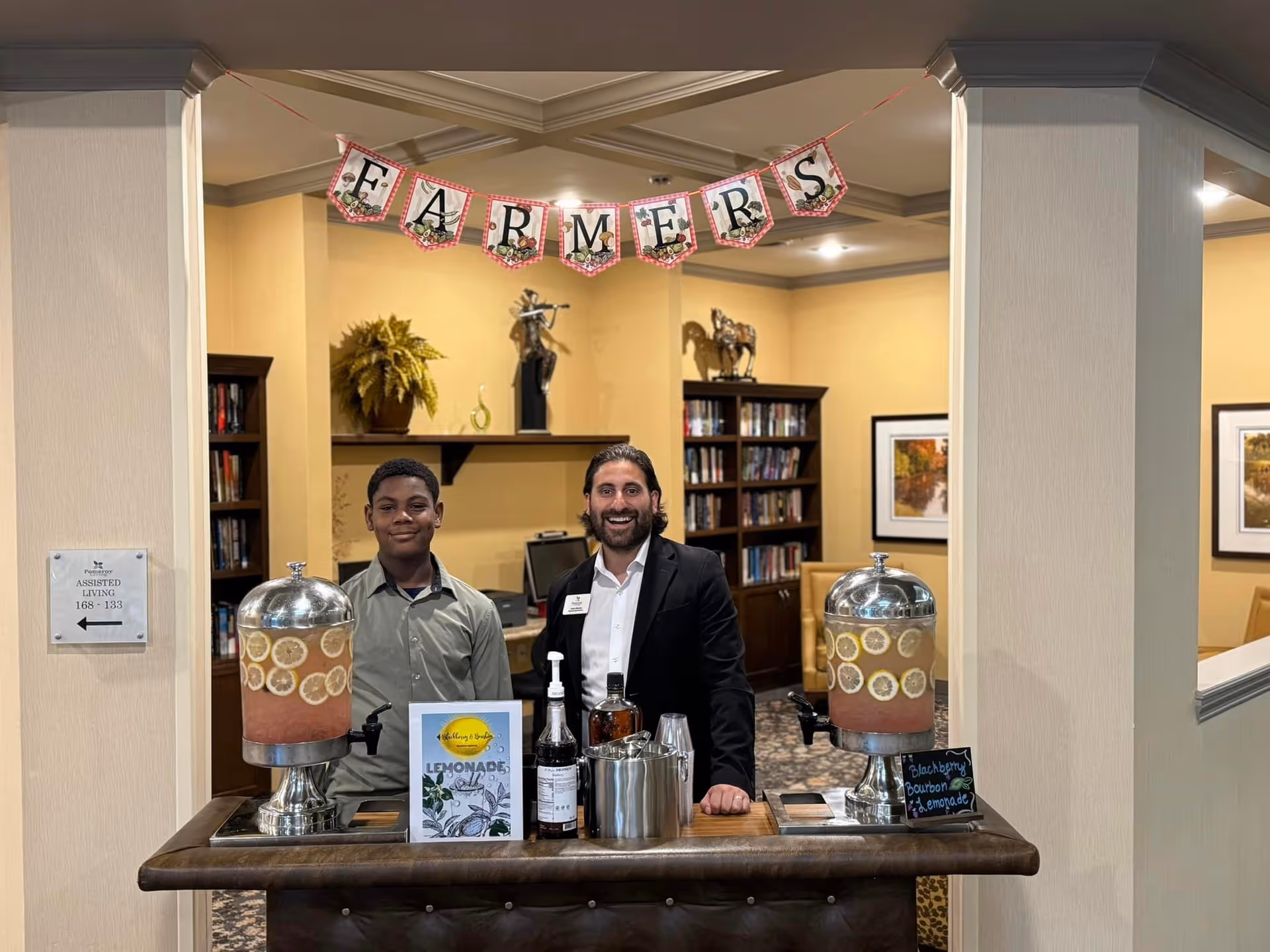 Two people stand behind a beverage cart with large lemonade dispensers in a decorated common room featuring bookshelves and a 'FARMERS' banner.