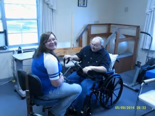 A woman sitting on a chair facing an elderly man in a wheelchair inside a room with exercise equipment and a window in the background.