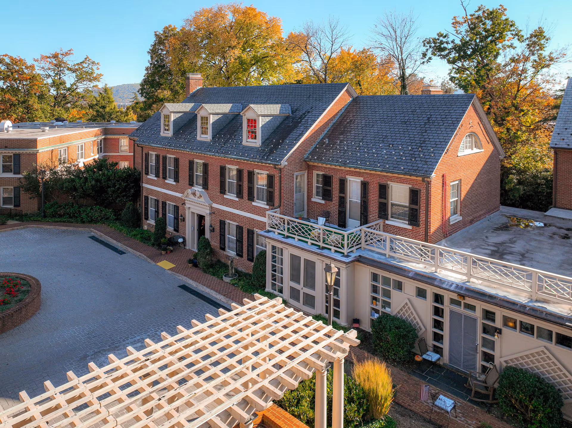 Exterior view of a brick senior living facility building with multiple windows and dormers on the roof, surrounded by trees with autumn foliage. There is a paved driveway with a circular flower bed and a white pergola structure in the foreground.
