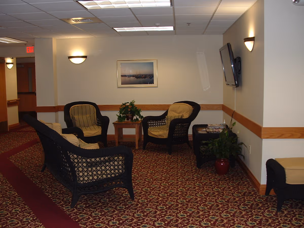 A cozy sitting area in a senior living facility with four cushioned wicker chairs arranged around a small wooden table with a potted plant. The room has patterned carpet, beige walls with wooden trim, wall-mounted lights, a framed picture on the wall, and a flat-screen TV mounted on the right wall. There is also a small side table with magazines and another potted plant on the floor.