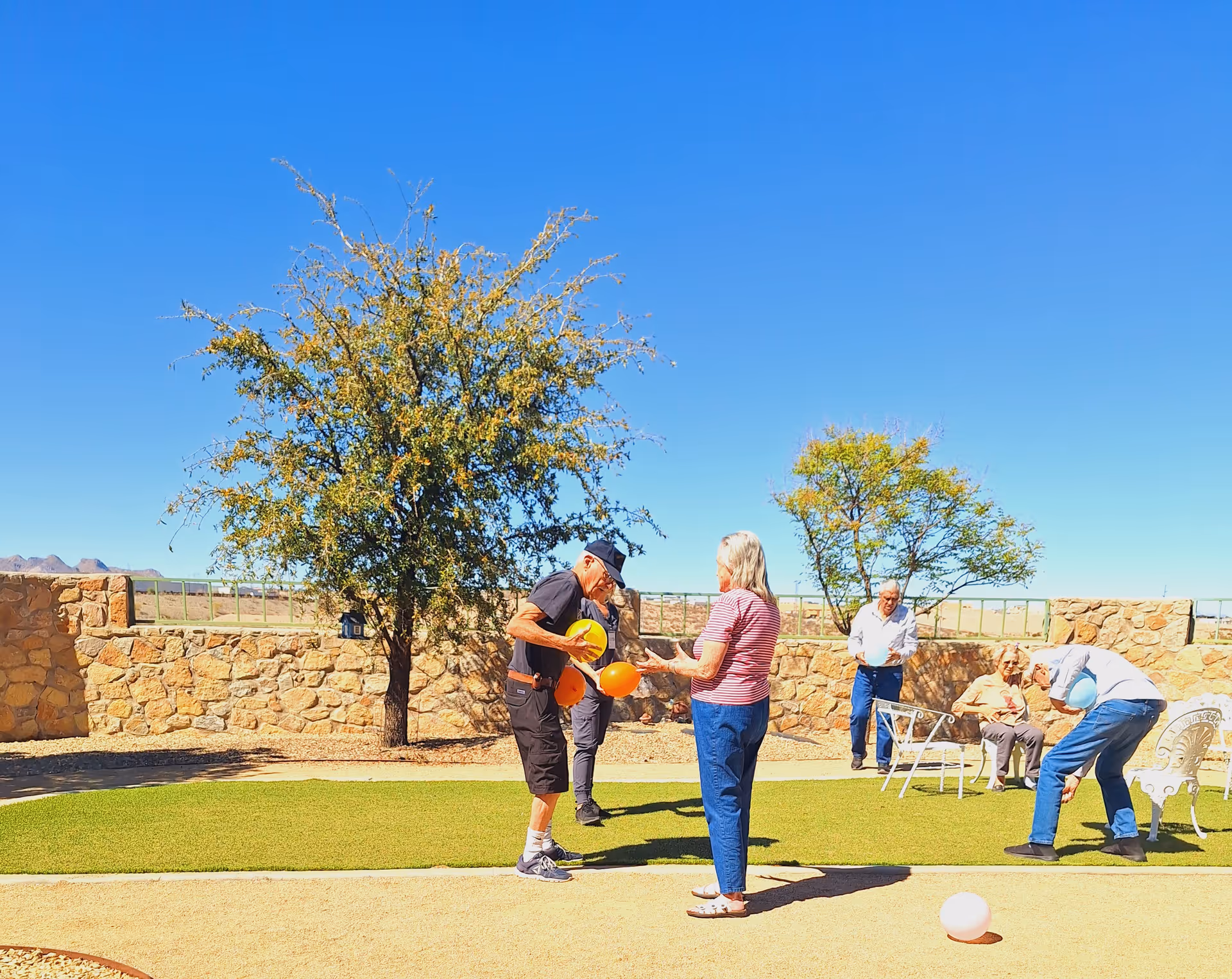 Seniors enjoying outdoor activities with balloons on a lawned courtyard bordered by stone walls and trees under a clear blue sky.