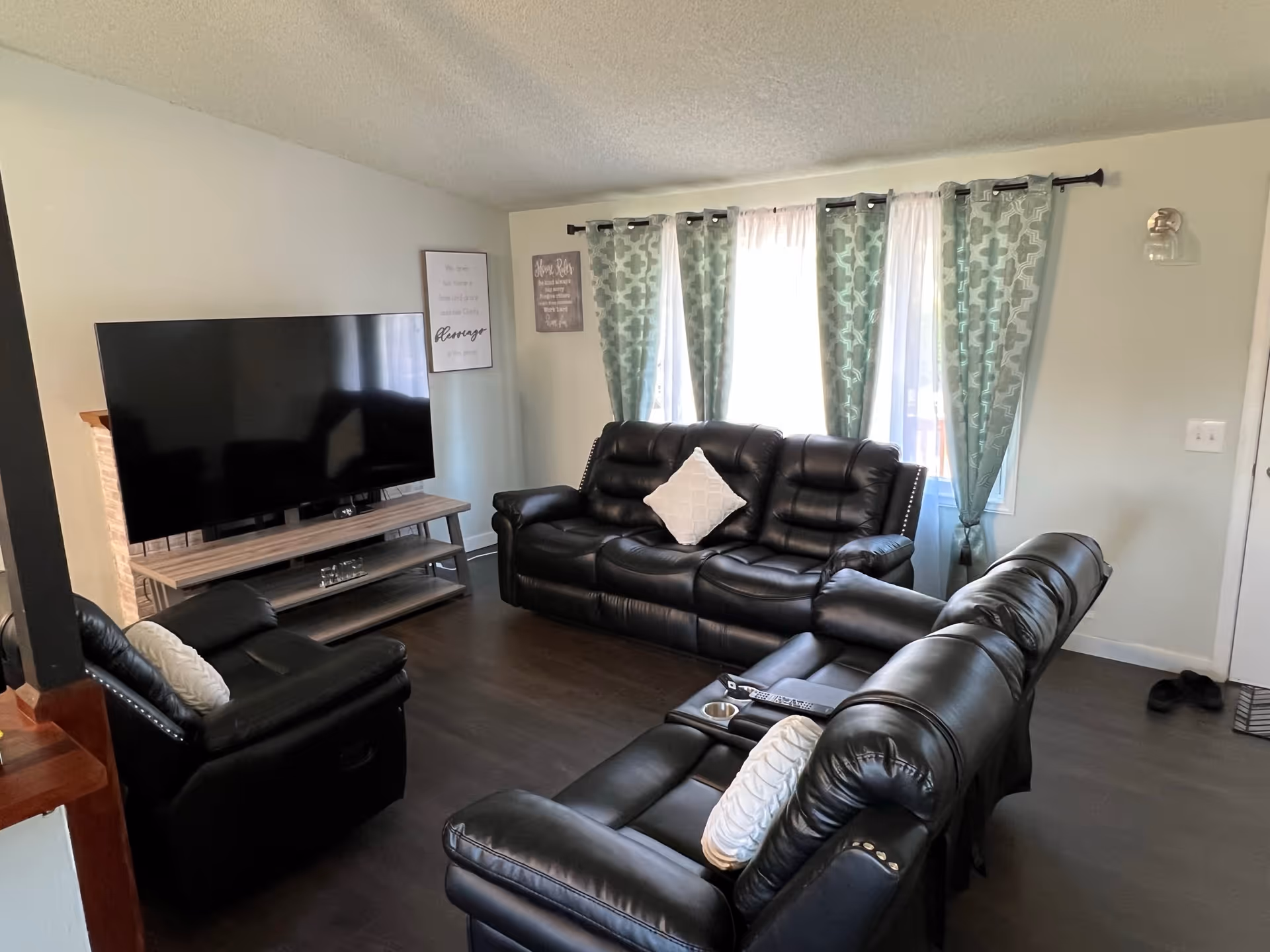 A cozy living room with black leather sofas and armchairs arranged around a large flat-screen TV on a wooden stand. The room has dark wood flooring, light-colored walls, and three windows with patterned green curtains letting in natural light. There are decorative pillows on the sofas and framed wall art near the TV.