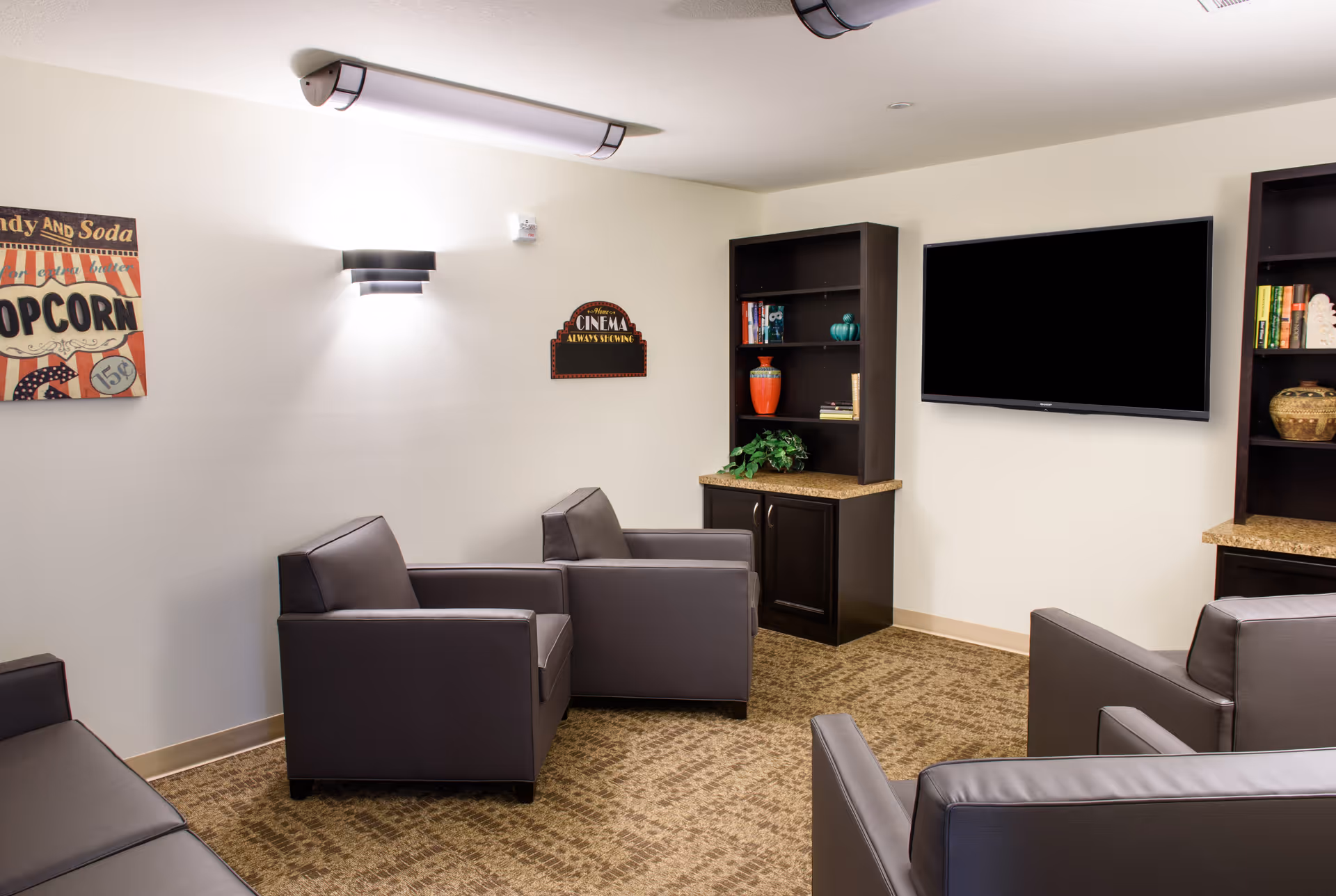 A cozy senior living common room with four gray armchairs arranged around a carpeted floor. The room features two dark wood bookshelves with decorative items and books, a flat-screen TV mounted on the wall, and two wall decorations including a popcorn sign and a cinema sign. The walls are light-colored, and the room is softly lit by ceiling and wall lights.