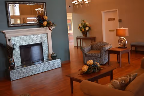 Cozy living room lounge with a tiled fireplace, armchairs, coffee tables with floral arrangements, and hardwood floors.