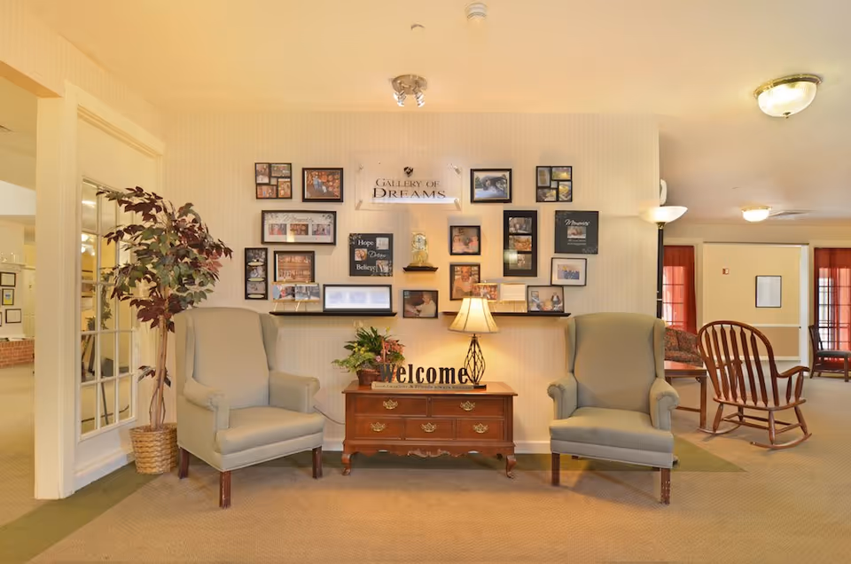 Welcoming sitting area with two upholstered armchairs flanking a wooden console table topped with a lamp and a 'Welcome' sign beneath a gallery wall of photos.