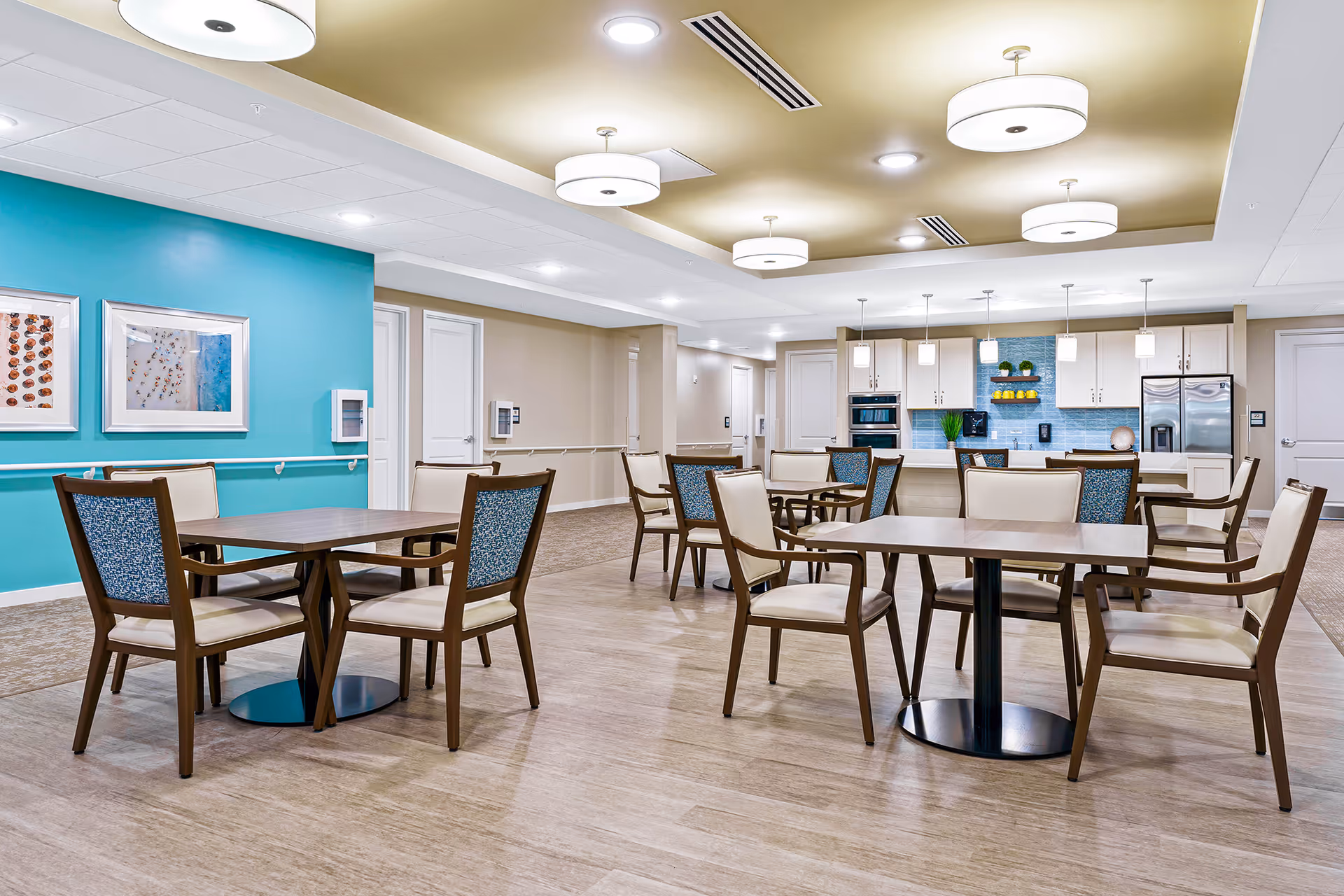 A bright and modern dining area in a senior living facility featuring multiple square wooden tables with cushioned chairs. The room has a light wood floor, a teal accent wall with framed pictures, and a kitchen area in the background with white cabinets, stainless steel appliances, and pendant lighting.