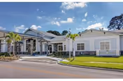 Exterior view of a single-story senior living facility building with a covered entrance, palm trees, and a well-maintained lawn under a partly cloudy sky.