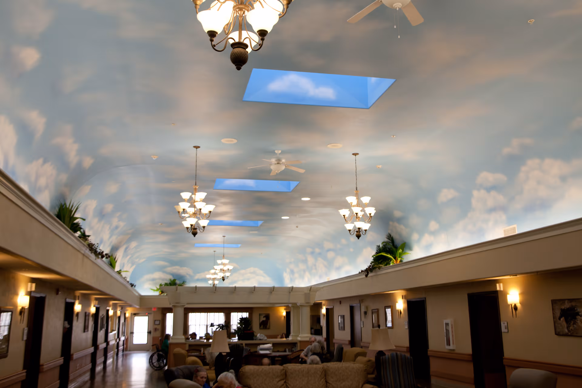Interior view of a senior living facility common area with a high ceiling painted to resemble a blue sky with white clouds. The ceiling has several skylights and hanging chandeliers. The room is furnished with sofas and chairs, and a few elderly people are seated. The walls have sconces and some plants are placed on ledges near the ceiling.