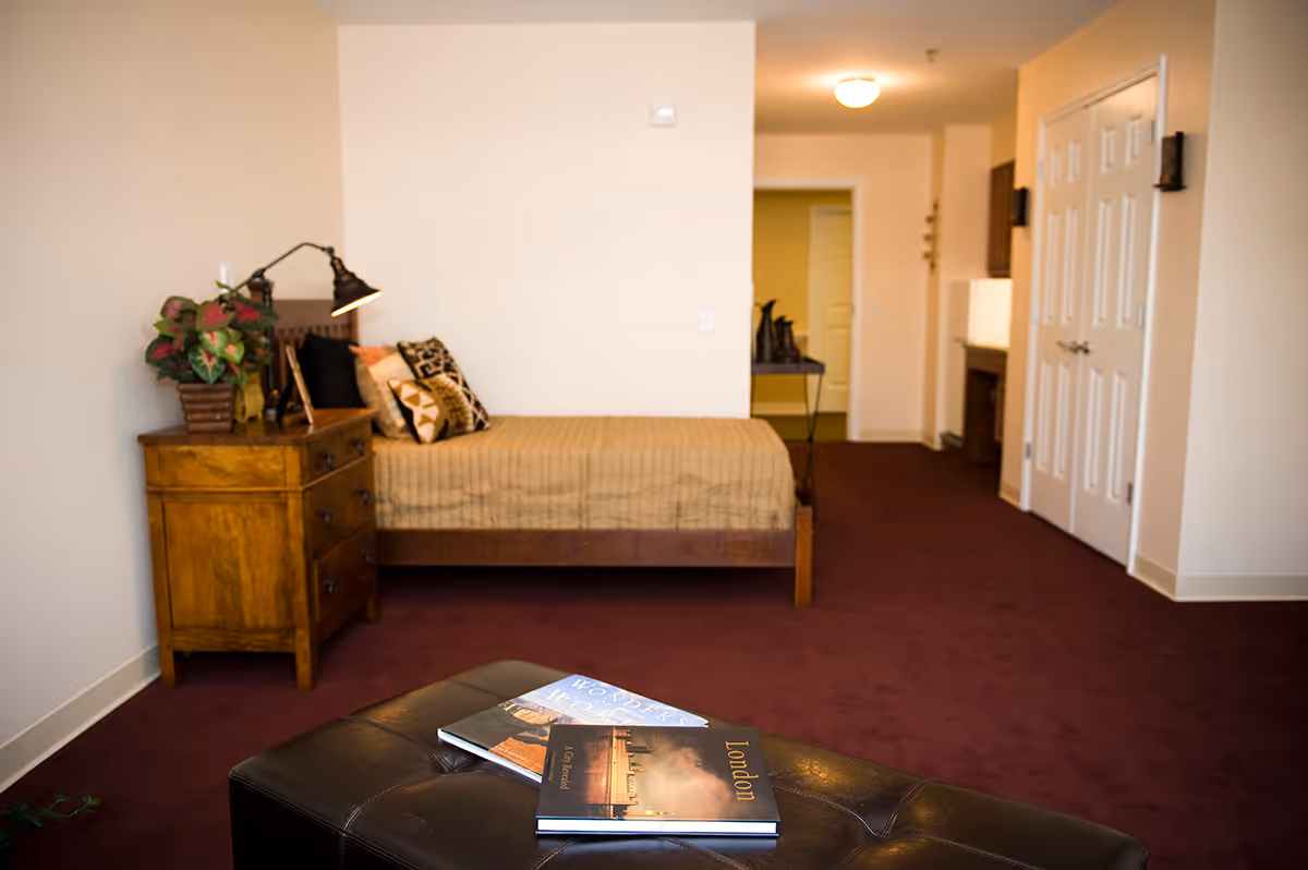 Studio-style interior showing a bed against the wall with a wooden nightstand and lamp, and an ottoman with books in the foreground.