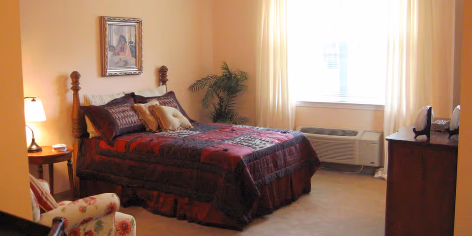 A cozy bedroom with a wooden bed frame, a bed covered with a red and dark patterned comforter and multiple pillows. There is a small wooden side table with a lamp and a cup, a floral upholstered armchair, a framed picture on the wall, a potted plant near the window, and a wooden dresser with decorative items. The room is softly lit with natural light coming through white curtains covering the window.