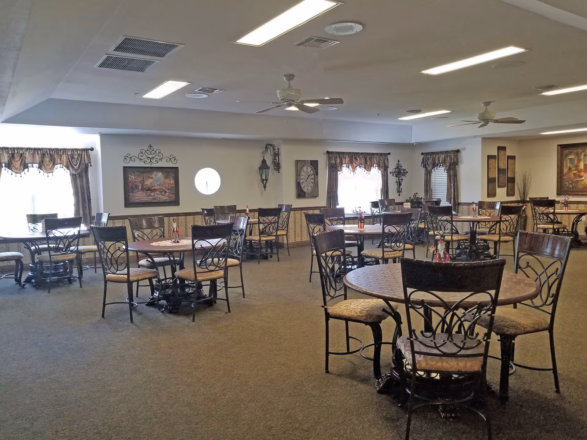 Spacious dining room with round tables and ornate chairs arranged under ceiling fans and fluorescent lights.