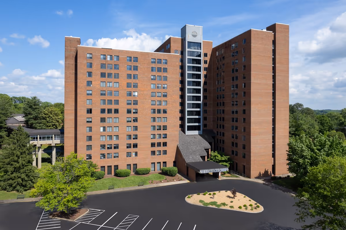 Large multi-story brick senior living building with a central entrance, parking area, and surrounding trees under a blue sky.