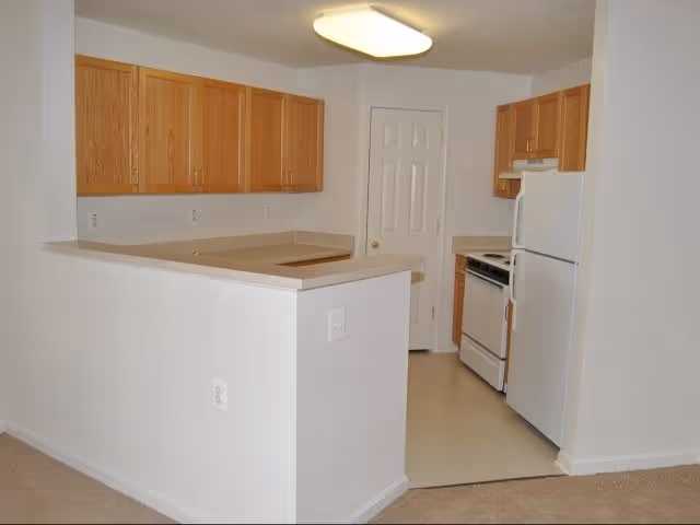 A small kitchen area in a senior living facility with light wood cabinets, white countertops, a white refrigerator, and a white stove. The kitchen is partially enclosed by a half wall and has a door at the back, likely a pantry or storage closet. The floor in the kitchen area is light-colored linoleum, and the adjacent area is carpeted.