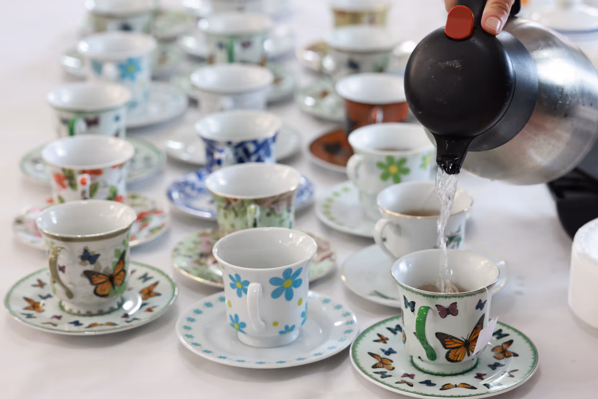 Multiple patterned teacups and saucers arranged on a table while a kettle pours hot water into one cup.