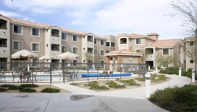 Outdoor courtyard area of Fountain View Village senior living facility featuring a fenced swimming pool with tables, chairs, and umbrellas, surrounded by a three-story beige building with tiled roofs under a partly cloudy sky.