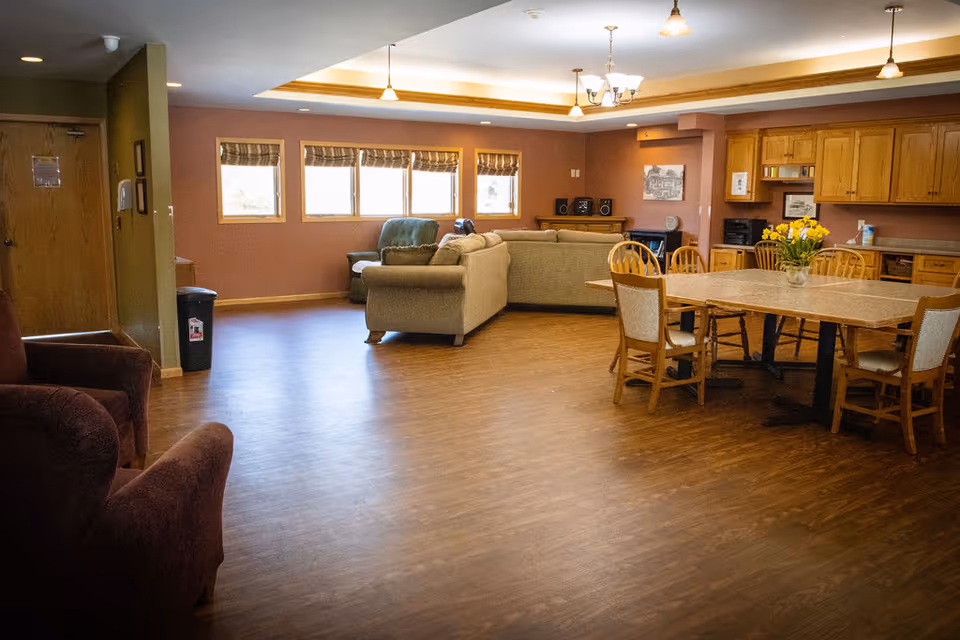 A spacious common area in a senior living facility featuring a wooden floor, a dining table with chairs, a comfortable seating area with sofas and armchairs, windows with blinds, and a kitchen area with wooden cabinets and a countertop. The room is well-lit with ceiling lights and has a warm, inviting atmosphere.