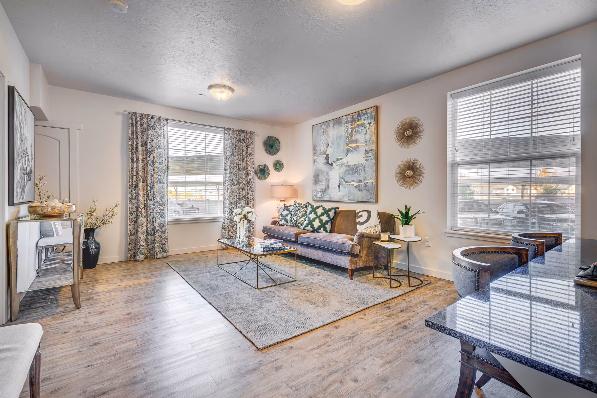 A bright and modern living room with a brown sofa adorned with patterned pillows, a glass coffee table with books and flowers, two large windows with blinds and patterned curtains, wooden flooring, and decorative wall art. There are also two small side tables with a lamp and a plant, and a granite countertop with three upholstered bar stools.