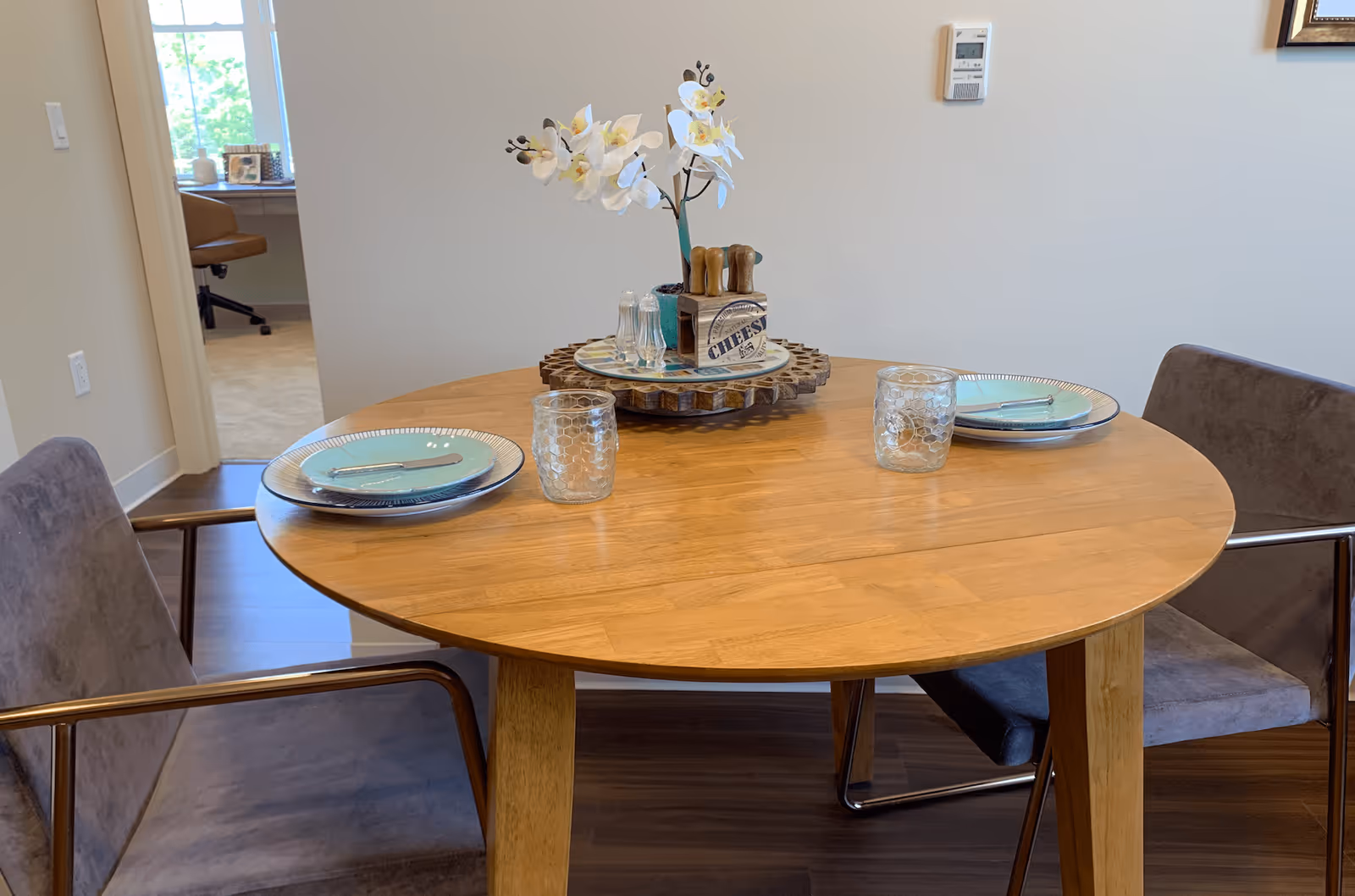Round wooden dining table set for two with plates, glasses, and a floral centerpiece in a dining area.