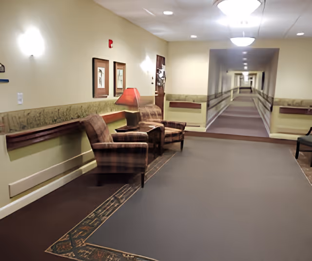 A long, well-lit hallway in a senior living facility with beige walls and carpeted floors. There are two upholstered armchairs with a small table and lamp between them on the left side, along with framed artwork on the wall. The hallway extends into the distance with handrails on both sides and additional seating areas visible.