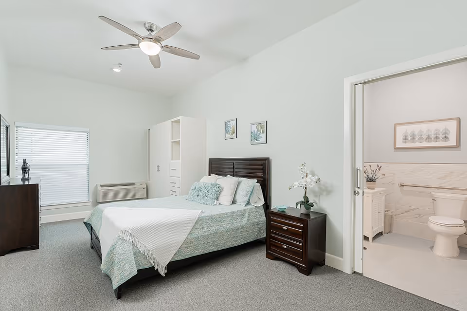 Well-lit furnished bedroom with a bed, nightstand, dresser, ceiling fan, and an open door revealing an adjoining bathroom.
