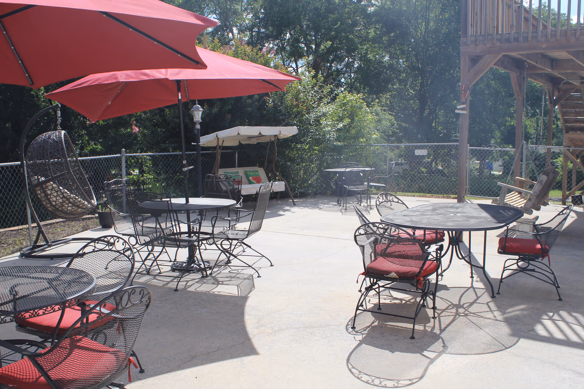 Outdoor patio area with several black metal tables and chairs with red cushions. Two large red umbrellas provide shade over some tables. There is a hanging wicker chair with a cushion, a wooden swing bench with a canopy, and a wooden staircase leading up to a deck. The area is enclosed by a chain-link fence with trees and greenery in the background.