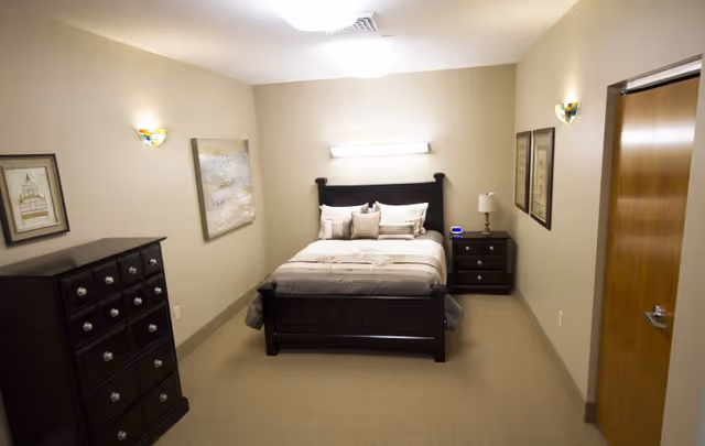 A neatly arranged bedroom with a dark wooden bed frame and a bed made with beige and white bedding. There are multiple pillows on the bed. On the left side of the room, there is a dark wooden dresser with several drawers. On the right side, there is a matching nightstand with a lamp and a small digital clock. The walls are beige with framed artwork hanging on them. The room is well-lit with ceiling lights and wall sconces. A closed wooden door is visible on the right side.