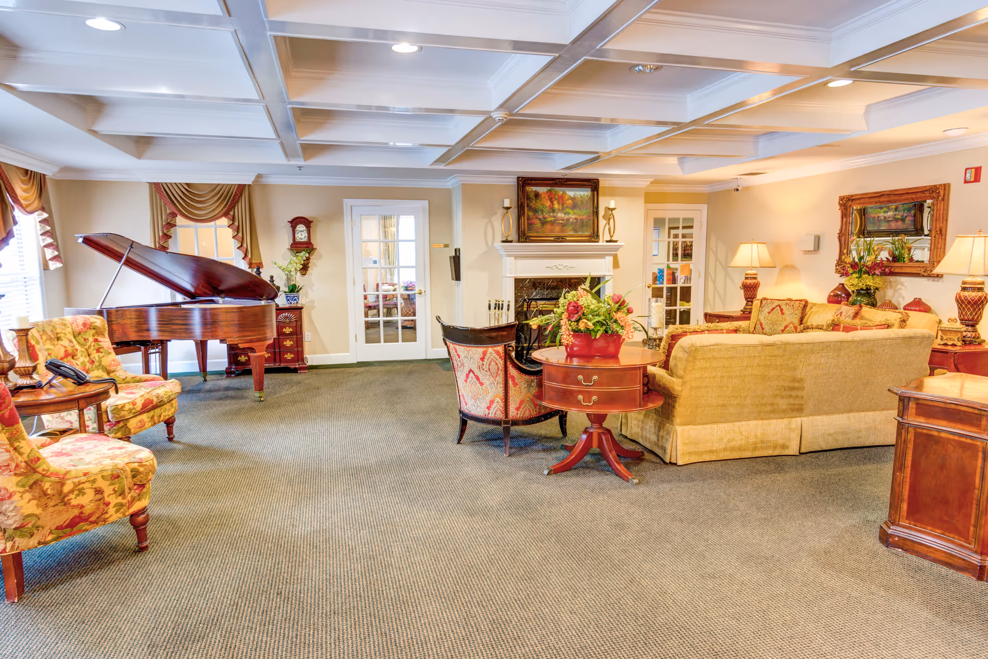 A spacious and elegantly decorated living room in a senior living facility featuring a grand piano, floral upholstered chairs, a beige sofa, a fireplace with a painting above it, and various tables with floral arrangements and lamps. The room has a coffered ceiling, carpeted floor, and large windows with draped curtains.