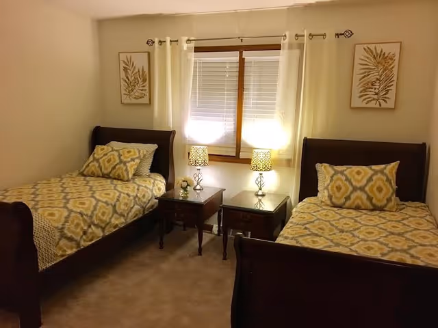 A bedroom with two twin beds featuring matching yellow and gray patterned bedding and pillows. Between the beds are two wooden nightstands with decorative lamps and a small flower arrangement. A window with white blinds and sheer curtains is centered on the wall behind the nightstands. Two framed botanical prints hang on the walls above each bed.