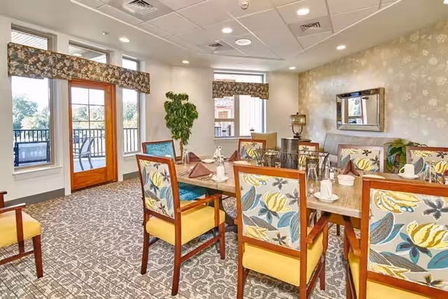 A bright dining room with a rectangular wooden table set for eight people. The chairs have wooden frames with yellow seats and floral-patterned backs. The room features large windows and a glass door leading to an outdoor patio with chairs. The walls are decorated with light-colored floral wallpaper and a large mirror. A green potted plant is placed near the windows.