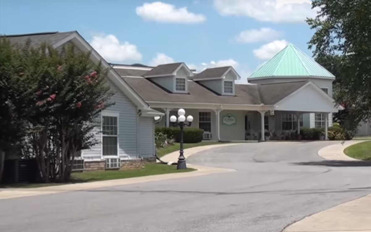 Exterior view of a senior living facility building with a light blue roofed tower section, white siding, and a covered entrance. There is a curved driveway leading to the entrance, a black lamp post with multiple white globes, and some green bushes and trees around the building under a partly cloudy sky.