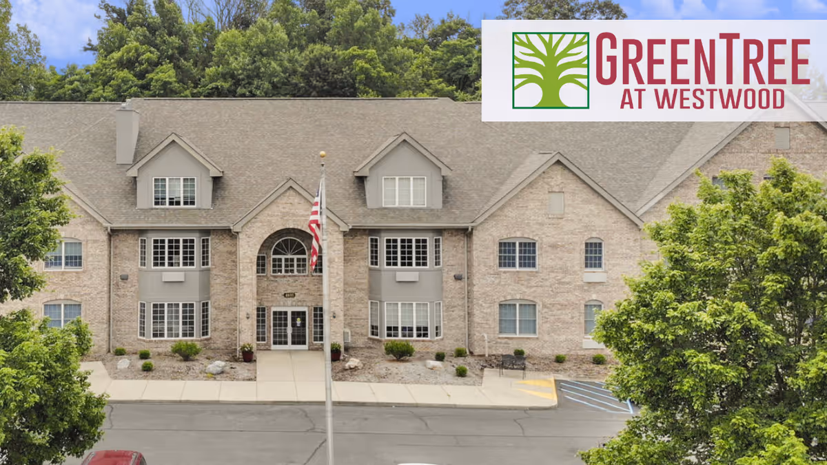 Exterior front view of a large brick building with multiple windows and a central entrance. There is a flagpole with an American flag in front of the building, surrounded by trees and greenery. The logo for GreenTree At Westwood is displayed in the top right corner.
