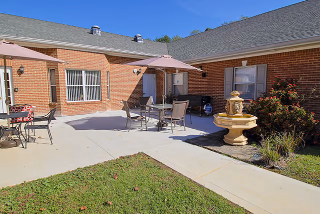Outdoor patio area at Sycamore Trace with brick building walls, several tables and chairs with umbrellas, a decorative stone fountain, and some greenery under a clear blue sky.