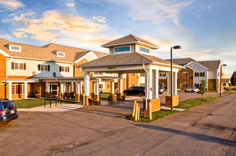 Exterior view of English Meadows Stephens City Campus, showing a large covered entrance with white columns and brick bases, surrounded by a multi-story building with beige siding and brick accents. Several cars are parked in the driveway and parking area under a partly cloudy sky during sunset.