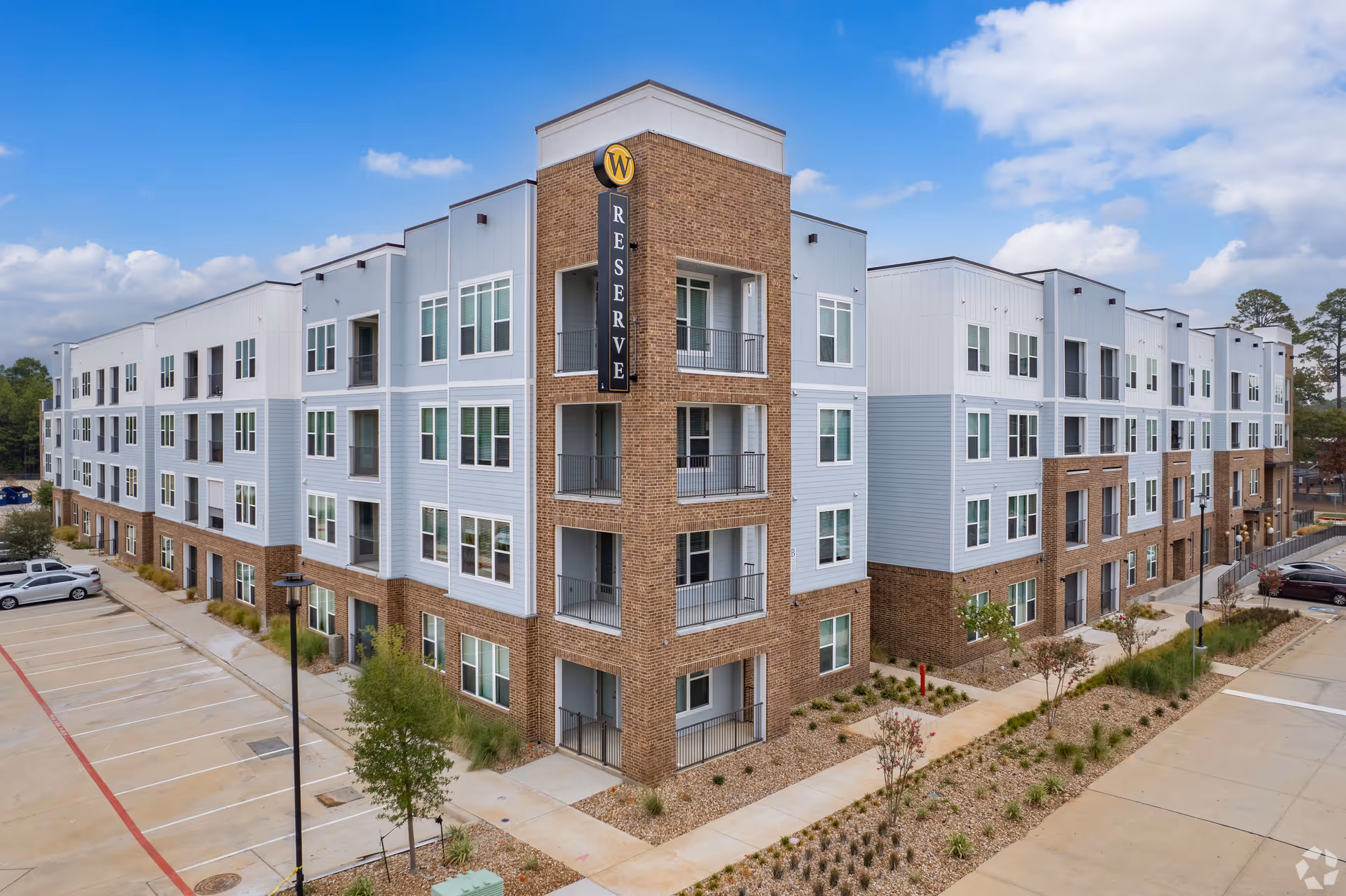Exterior view of a modern multi-story residential building with a combination of brick and light-colored siding. The building has multiple windows and balconies, with a sign that reads 'Reserve' on the corner. There is a parking lot with several cars and landscaped areas with small trees and shrubs surrounding the building under a partly cloudy sky.