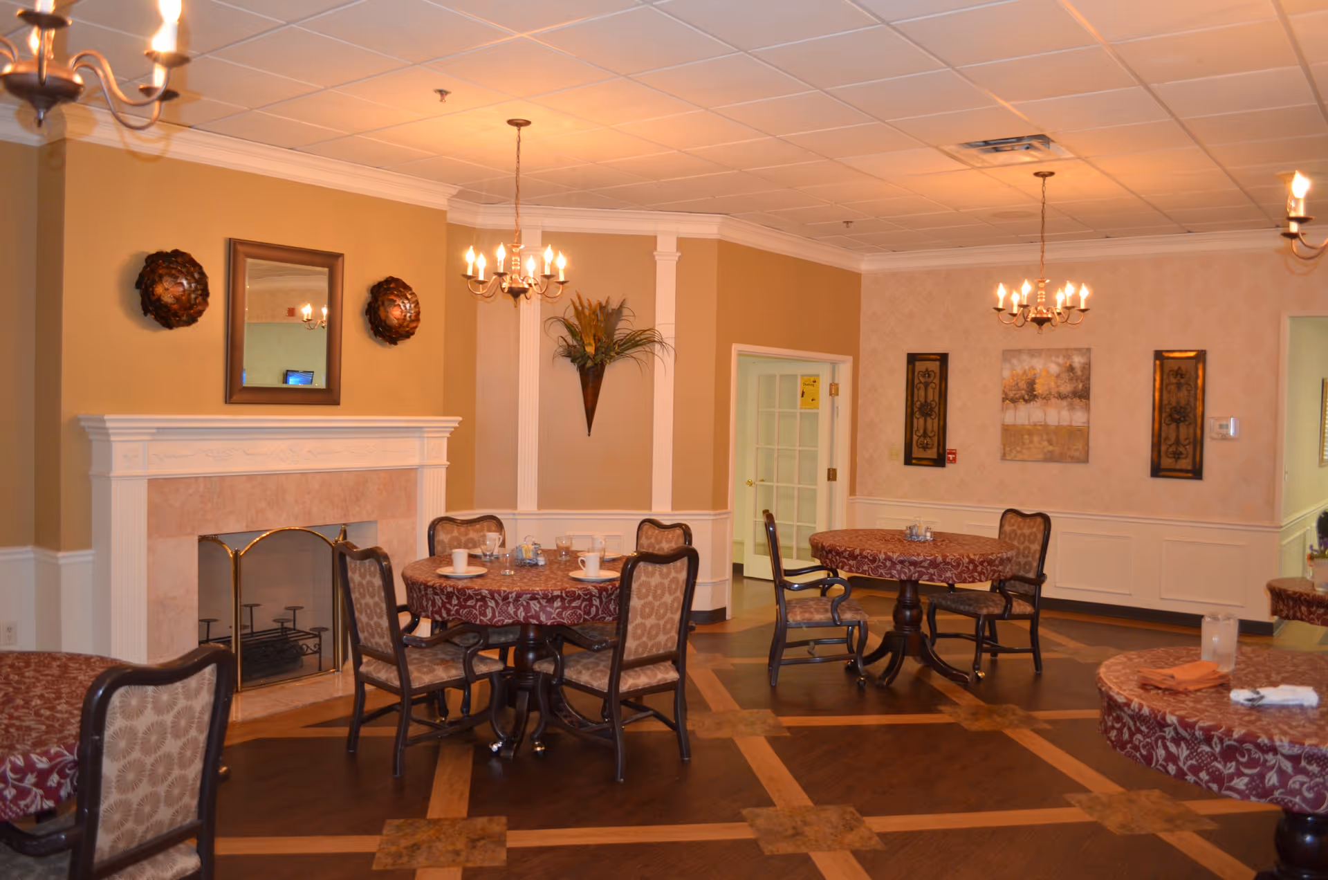 Cozy dining room with round tables and chairs, a fireplace, chandeliers, and warm beige decor.