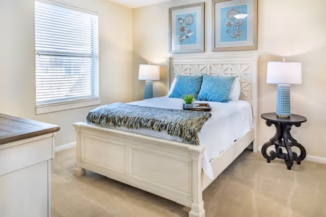 Bright bedroom with a white wooden bed dressed in blue pillows and a throw, flanked by matching lamps and framed artwork.