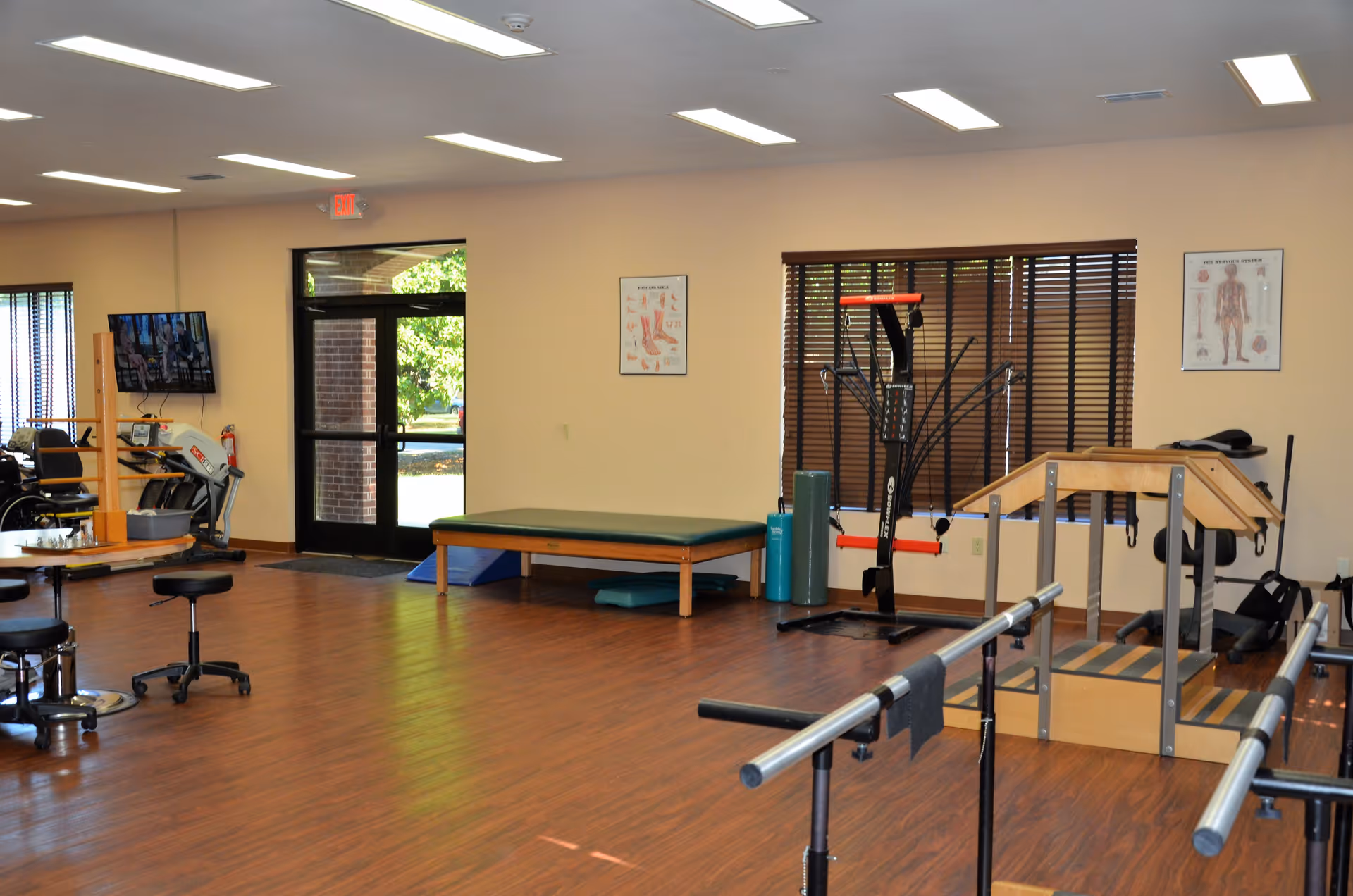 A spacious rehabilitation room with wooden flooring, featuring parallel bars for walking exercises, a padded therapy table, exercise equipment, anatomical charts on the walls, large windows with blinds, and a glass door leading outside.