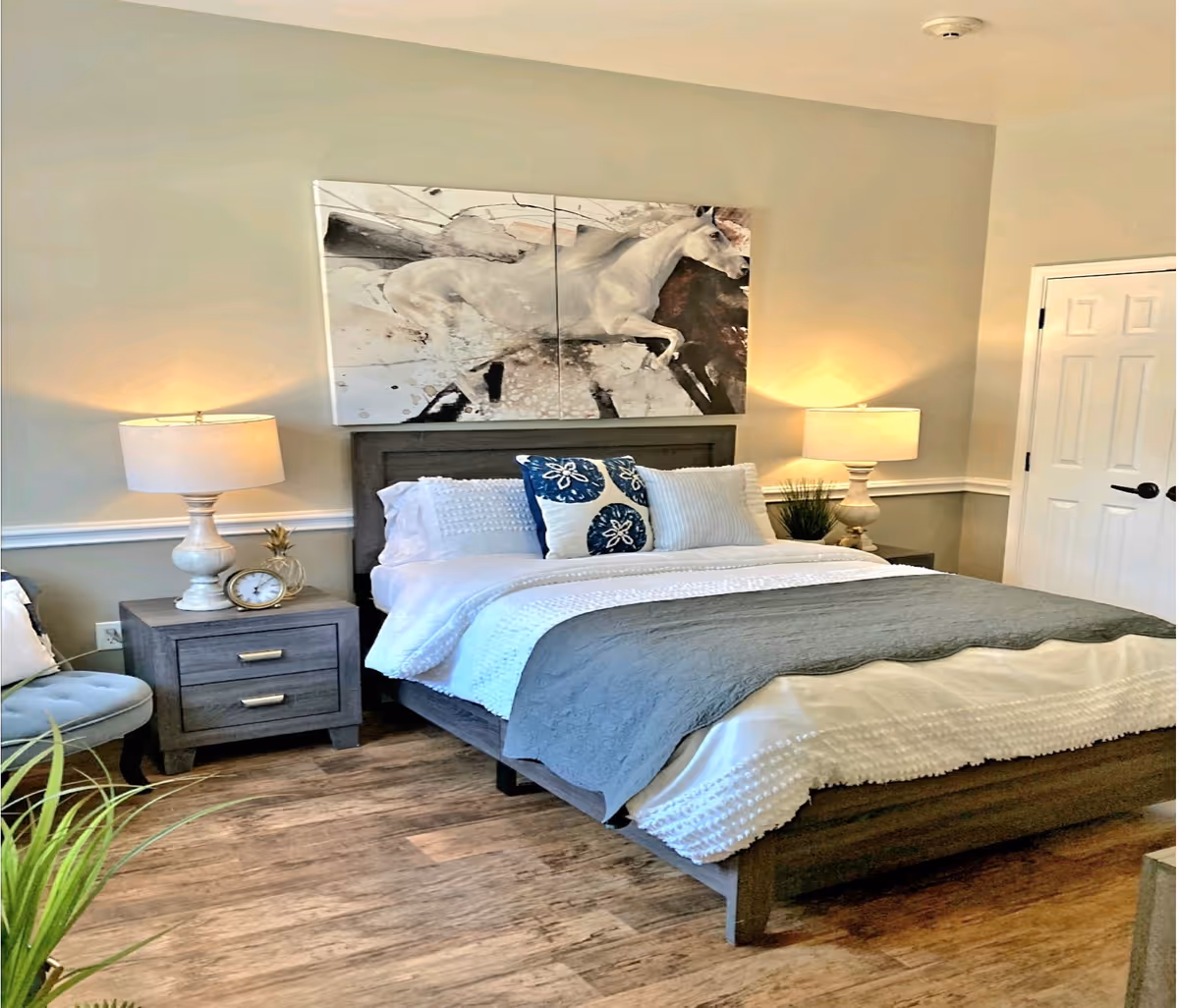 A neatly made bed with white and gray bedding and decorative pillows in a bedroom. Two bedside tables with lamps flank the bed. Above the bed is a large artwork of a white horse. The room has wooden flooring, a closed white door, and a light gray wall.