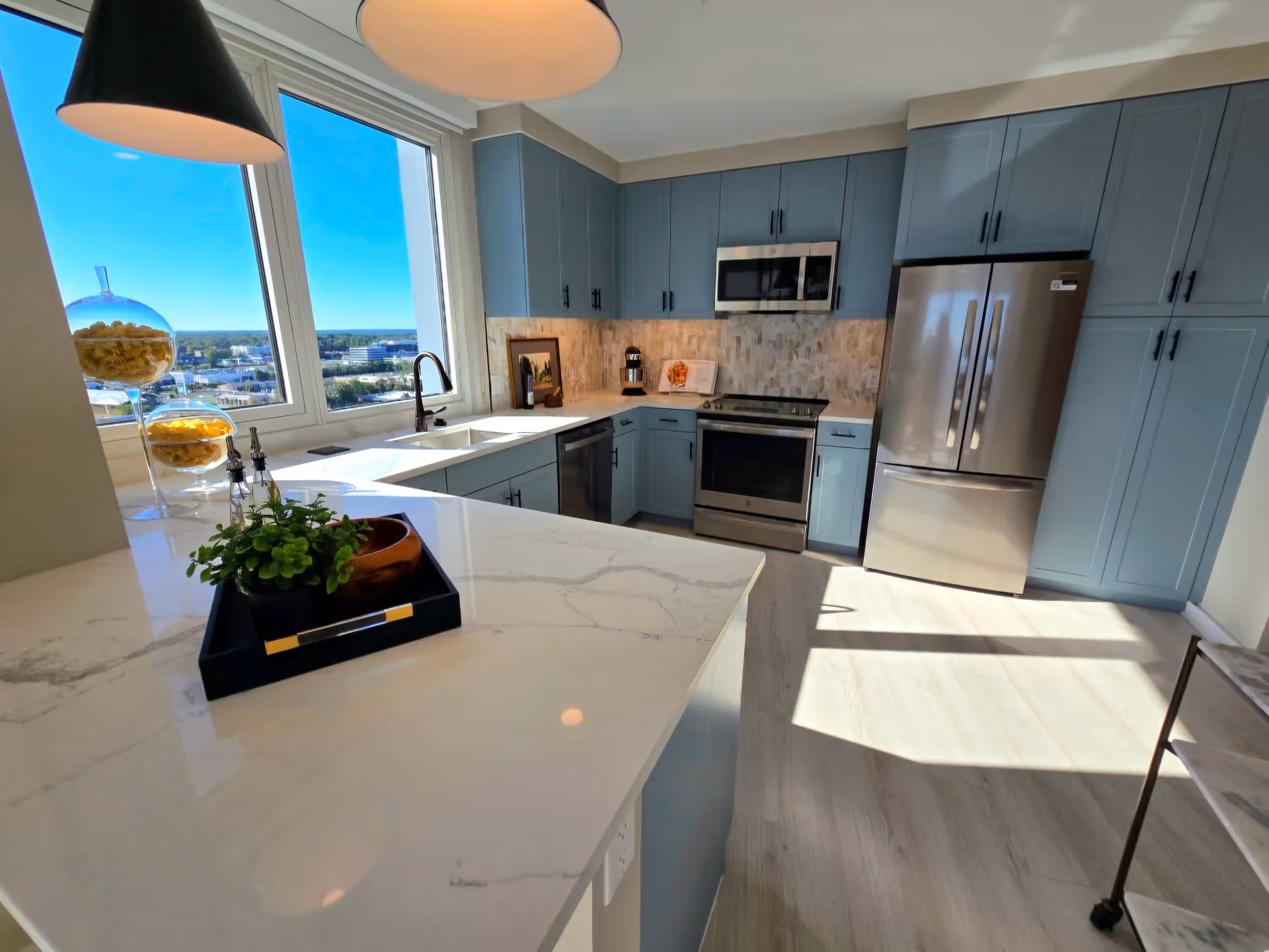Bright modern kitchen with light blue cabinets, stainless steel refrigerator, oven, and microwave. White marble countertops and a large window letting in natural light. A small plant and decorative items are on the counter.