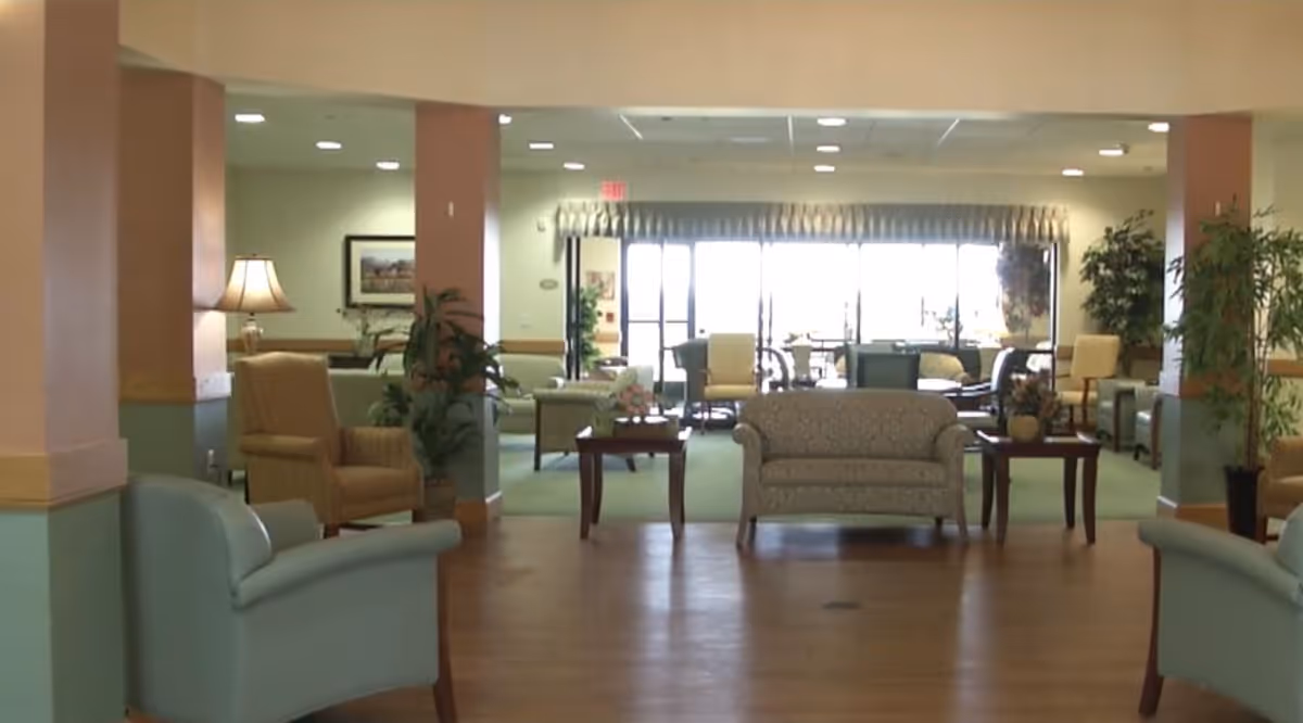 A spacious senior living facility common area with multiple armchairs and sofas arranged around small tables. The room has soft lighting, plants, and large windows in the background letting in natural light.