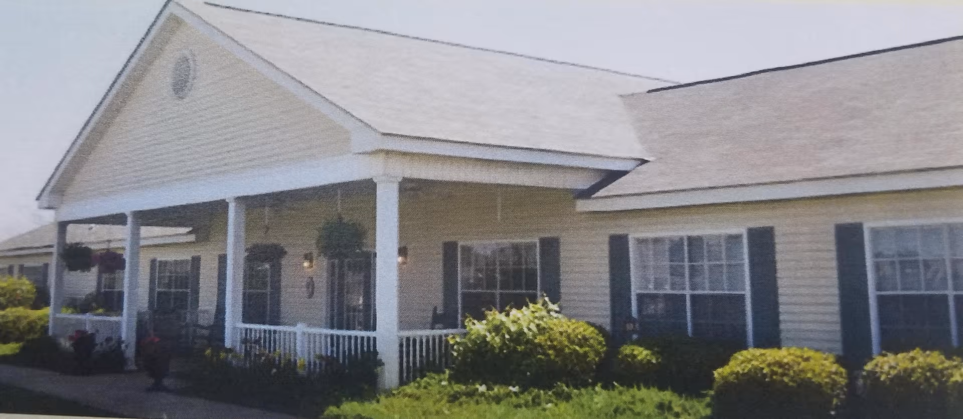 Single-story senior living building with a covered porch, white columns, hanging plants, and landscaped shrubs.