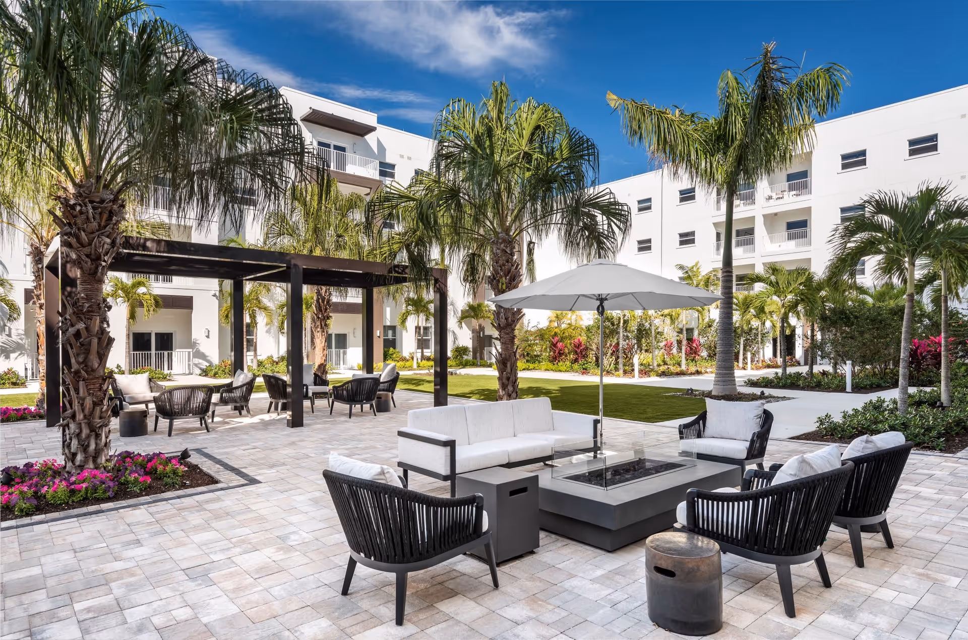 Outdoor courtyard area at Amavida at Lakes Park featuring modern patio furniture including cushioned chairs and a sofa arranged around a rectangular fire pit table with a large white umbrella. The space is paved with light-colored tiles and surrounded by palm trees, colorful flowers, and a well-maintained lawn. White multi-story buildings with balconies are visible in the background under a blue sky with some clouds.