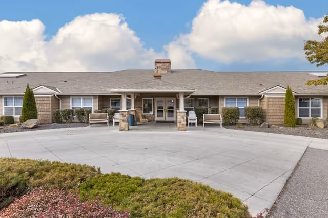 Front exterior view of a single-story senior living facility building with a covered entrance supported by stone pillars, benches on either side, and landscaped bushes and trees under a partly cloudy sky.