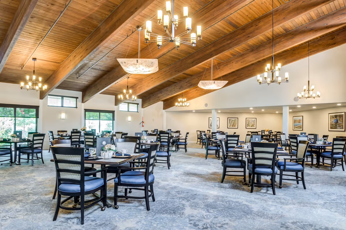 Spacious dining room with multiple round tables and black chairs with blue cushions. The room features a high wooden ceiling with exposed beams and several modern chandeliers. Large windows allow natural light to brighten the space, and framed artwork decorates the walls.