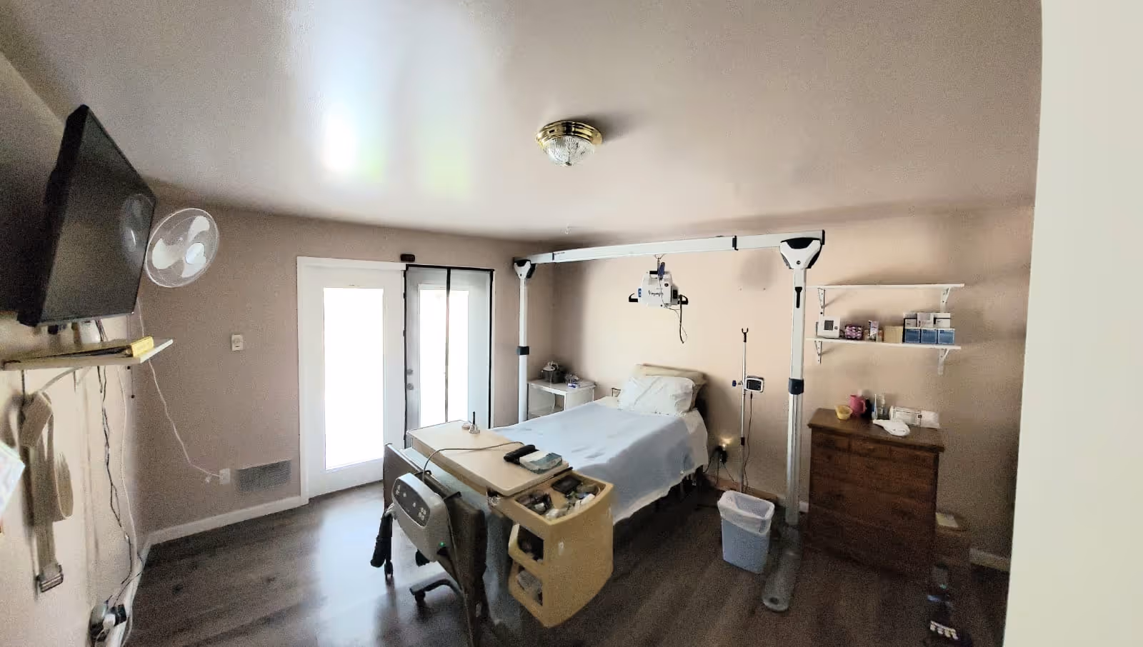 A small, well-lit room with a hospital bed equipped with medical devices including an overhead patient lift. The room has light-colored walls, a wooden floor, a wall-mounted TV, a fan, a dresser with medical supplies, and a door with glass panels letting in natural light.