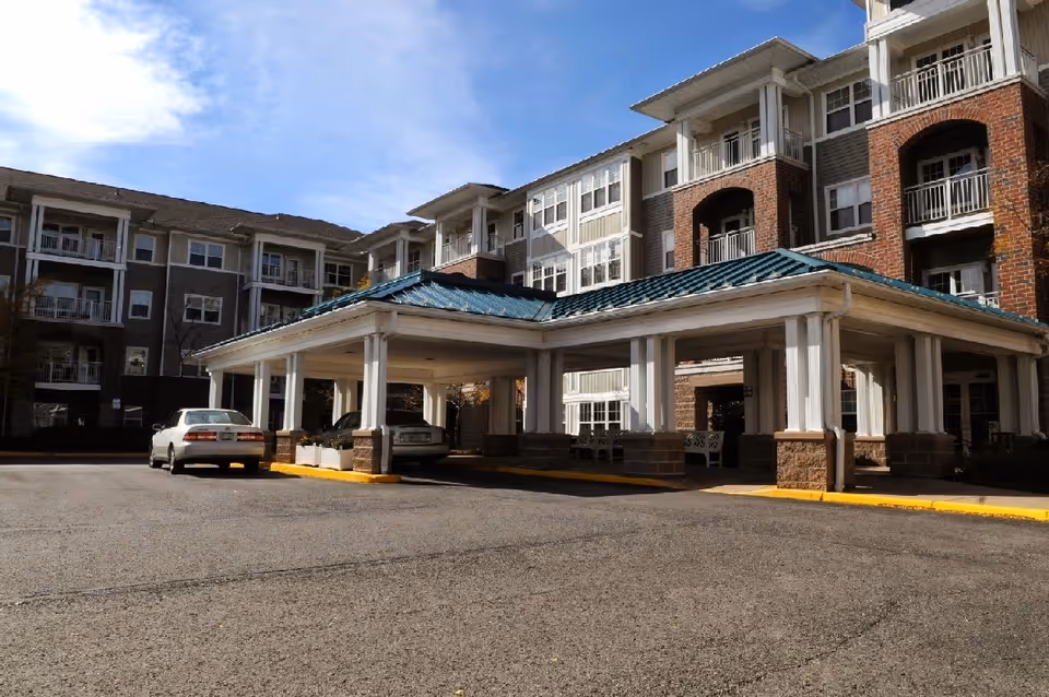 Exterior view of a multi-story senior living facility with a covered entrance area and parked cars in front under a blue sky.