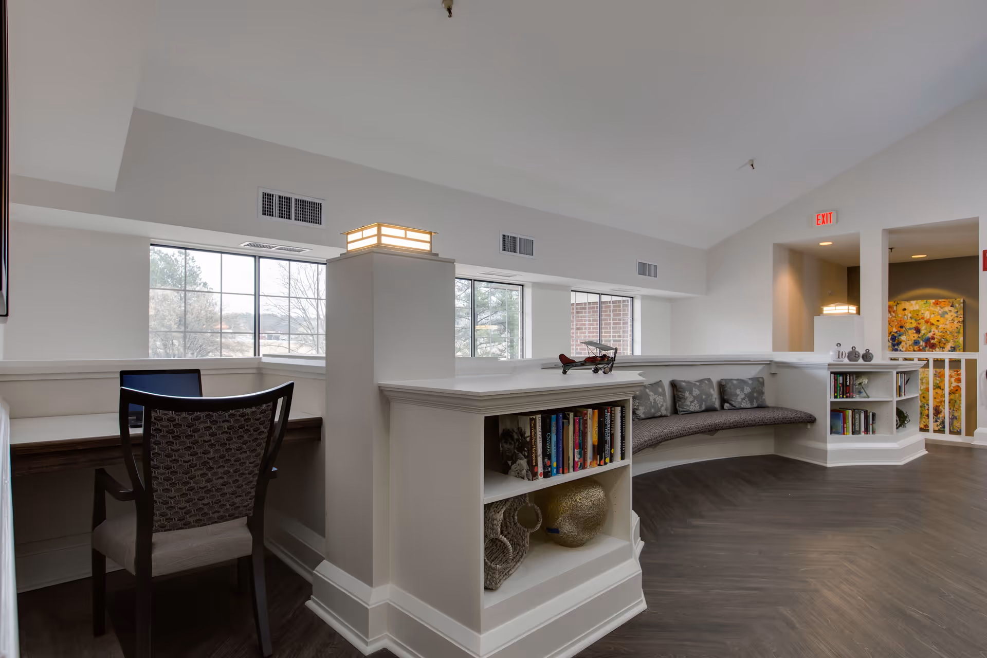 Bright communal lounge with built-in bookshelves, a curved cushioned bench, and a desk and chair by large windows.