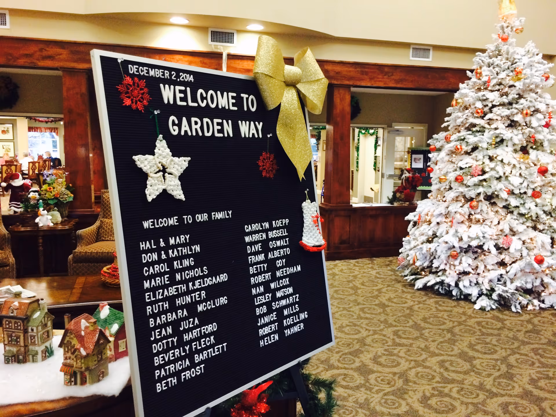 Interior of Garden Way Retirement Community decorated for Christmas with a large white Christmas tree adorned with red ornaments on the right. In the foreground, a black letter board with white letters welcomes residents and lists their names, decorated with a large gold bow, a white star, and a white bell ornament. The background shows a cozy seating area with chairs and holiday decorations.