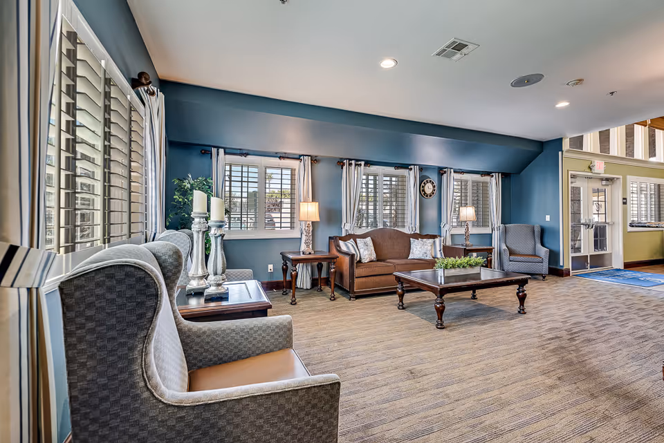 Spacious lounge area with upholstered armchairs and a sofa arranged around a wooden coffee table beneath windows with shutters.