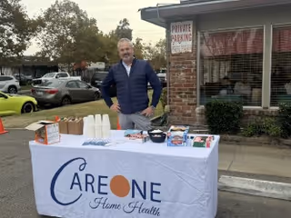 A man stands behind a table covered with a white tablecloth displaying the CareOne Home Health logo. The table has various items including cups, snacks, and informational materials. The setting is outdoors near a brick building with a 'Private Parking' sign visible, and several parked cars and trees are in the background.